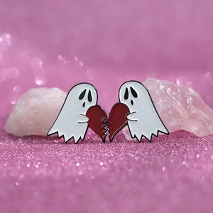 Detail of two cartoon ghost enamel pins holding a broken red heart on pink glitter