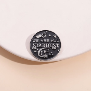 Macro shot of circular enamel pin featuring stars and We Are All Stardust text engraving