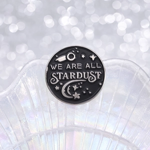 Black round enamel badge pin with We Are All Stardust text and celestial star patterns