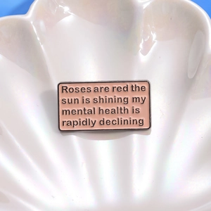 Close-up of a pink enamel pin with black humorous text on a white shell-shaped dish