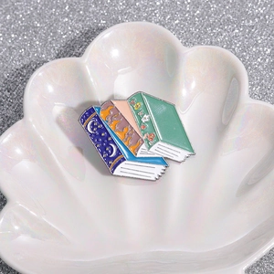 Detailed close-up of three stacked book enamel pins on a decorative shell tray