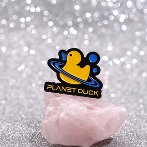 Détail du pin's émaillé Planet Duck jaune avec anneau bleu sur une pierre de cristal rose