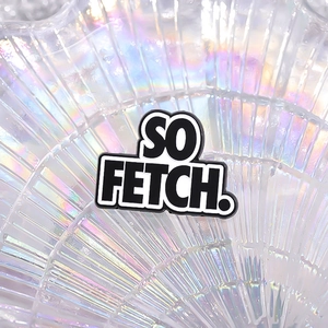 Close-up of 'SO FETCH' black and white enamel pin on an iridescent pearl tray