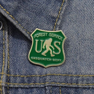 Green enamel Sasquatch Forest Service shield brooch pinned on blue denim fabric