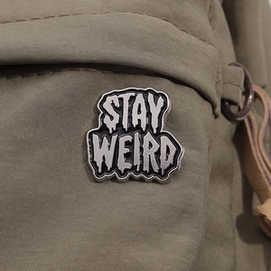 Macro shot of 'STAY WEIRD' black enamel pin on an olive green canvas bag