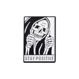 Detail of enamel pin featuring skull in cloak with thumbs up and Stay Positive text