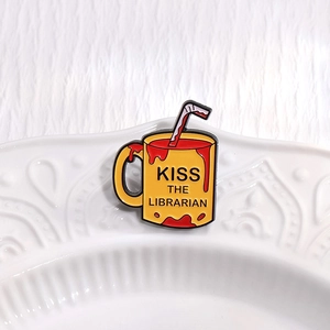 Close-up of yellow mug enamel pin with red liquid and Kiss The Librarian text on ceramic