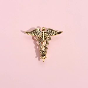 Golden medical caduceus brooch with winged snakes and staff on a soft pink background