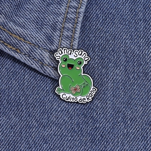 Detail of green enamel frog pin with Sana sana Culito de Rana text on denim fabric