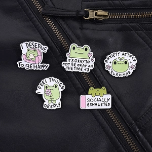 Detail view of five cartoon frog enamel pins with empowering slogans on black leather