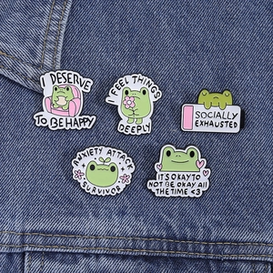 Set of 5 cute green frog enamel pins with motivational quotes on blue denim background
