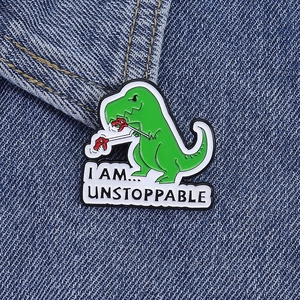 Dettaglio della spilla in smalto T-Rex verde con slogan UNSTOPPABLE su denim blu