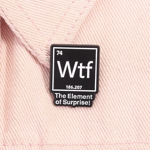 Macro d'un pin's chimie en émail noir avec texte 'Element of Surprise' sur denim rose