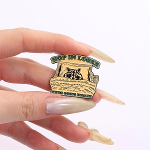 Macro detail of enamel pin with raccoon in bed and 'Hop In Loser' humorous text