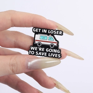 Macro shot of ambulance enamel pin with 'Get in loser we're going to save lives' text