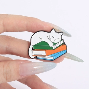 Detail of cute cat sleeping on books enamel pin held in hand to show the actual product size