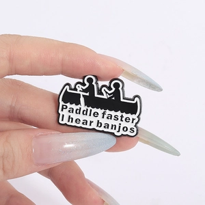 Close-up of hand holding black enamel pin with Paddle Faster I Hear Banjos text