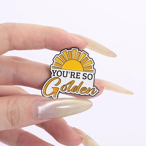 Macro view of 'YOU'RE SO Golden' sun enamel pin badge held in hand against white background