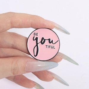 Gros plan d'une main tenant une broche ronde en émail rose avec l'inscription BE YOU TIFUL