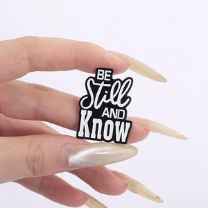 Detailed shot of black enamel metal pin with white BE STILL AND KNOW lettering design