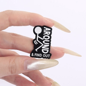 Detail of hand holding a black enamel pin with white text and stick figure graphics