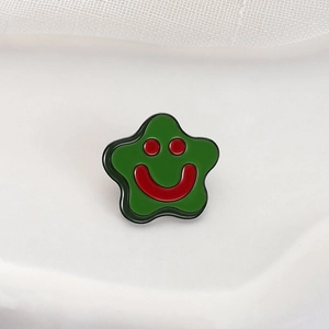 Cartoon star-shaped metal enamel pin with green base and red smiling face on white fabric