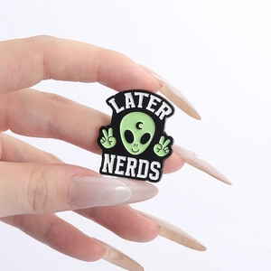 Dettaglio di spilla smaltata aliena verde con simbolo della luna e testo Later Nerds