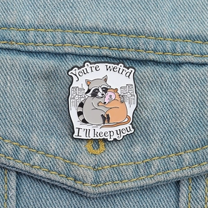Cute raccoon and opossum enamel pin badge on denim jacket with You're Weird quote