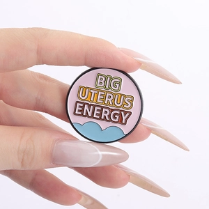 Detail of pink enamel pin with Big Uterus Energy text held by hand on white background
