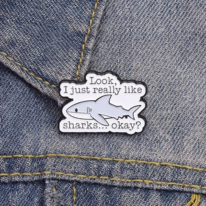 Cartoon shark enamel pin with humorous text displayed on a denim jacket lapel