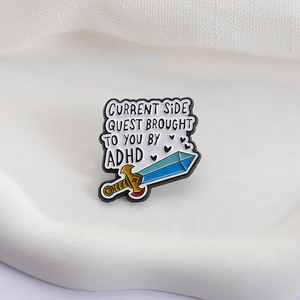 Enamel sword pin with ADHD side quest quote on white textured ceramic tray