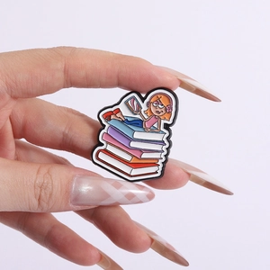 Detailed view of girl reading on stacked books enamel pin held by hand to show size
