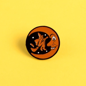 Halloween enamel pin featuring an orange moon face and silhouette of a witch on a broomstick