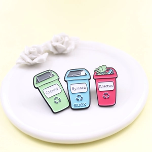 Three colorful enamel trash bin pins with recycling symbols on white ceramic tray
