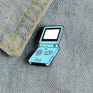 Hellblaue Retro-Klappspielkonsole-Emaille-Anstecknadel auf blauem Denim-Hintergrund