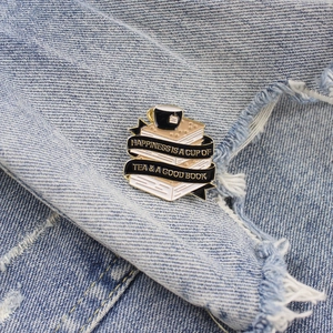Detail of Happiness book and tea enamel pin on denim fabric with gold metal finish