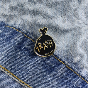 Creative black trash bag enamel pin with gold Trash text on denim fabric background