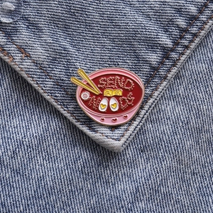 Creative enamel pin featuring a ramen bowl with a heart egg and SEND NOODS text