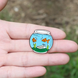 Close-up of a creative round fish tank enamel pin featuring orange goldfish and a grey bed