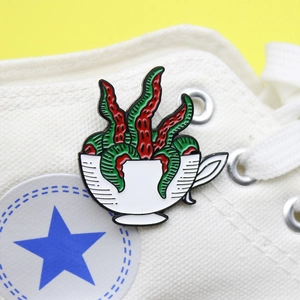 Detailed enamel pin featuring red and green tentacles in a white teacup on canvas