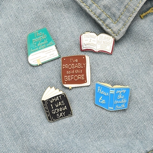 Five creative book-shaped enamel pins in various colors on blue denim fabric background