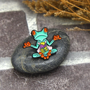 Green zen frog enamel pin with colorful anatomical design on a dark pebble stone