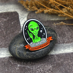 Alien in silver UFO enamel pin with Greetings from Aliens text on dark pebble stone
