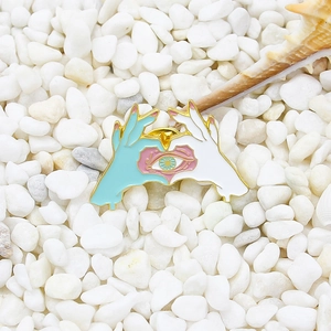 Artistic heart hands enamel brooch with sunburst eye motif on white pebbles background