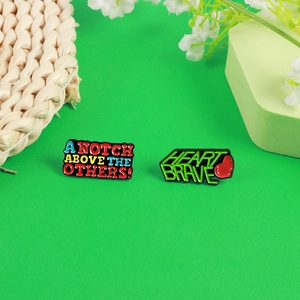 Enamel pins featuring 'A Notch Above The Others' and 'Heart Brave' slogans on green background