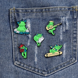 Set of five cute cartoon frog enamel pins displayed on a denim pocket accessory
