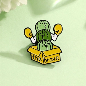 Macro shot of cartoon cactus enamel pin in a yellow box with 'the brave' text