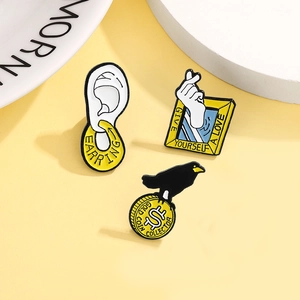 Three creative enamel pins including heart gesture, ear shape, and crow on gold coin