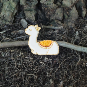 Close-up of a cute white alpaca enamel brooch with colorful saddle on tree bark