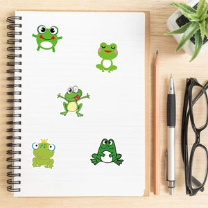 Five cute cartoon frog stickers displayed on a notebook page with wooden background